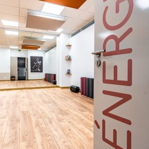 Studio de fitness moderne avec parquet en bois clair, miroir mural, et équipements de yoga soigneusement rangés, créant une ambiance dynamique et accueillante. La porte entrouverte marquée "ÉNERGIE" souligne l'atmosphère active du lieu.